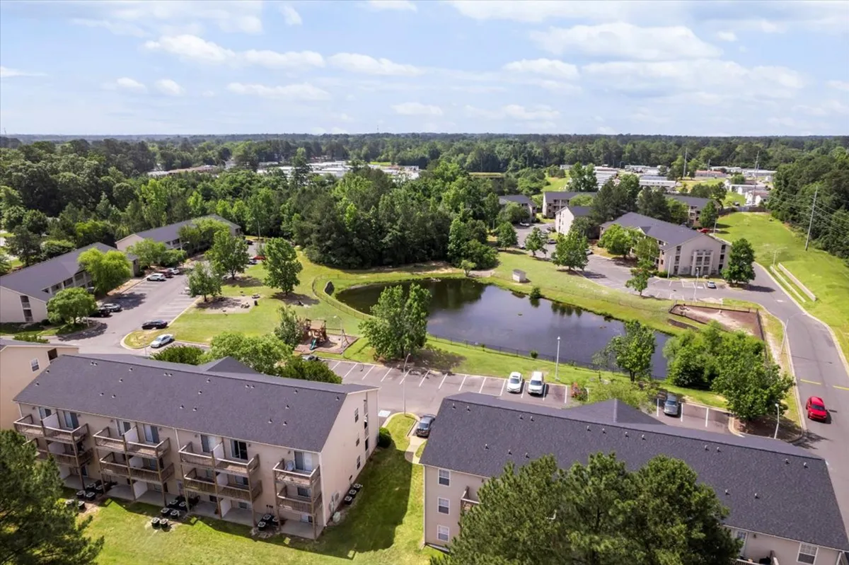 A peaceful pond framed by green lawns and trees, with apartment buildings in the background. Residents enjoy walking paths, picnics, or simply relaxing by the water, hinting at a slower pace and natural beauty within the community.