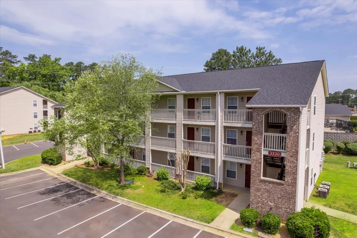 A three-story apartment building with well-maintained green landscaping and a spacious parking area. The scene is peaceful, showing plenty of room for residents and suggesting ease of coming and going, fitting for those with demanding, on-the-go schedules.