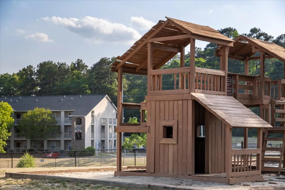 A lively outdoor scene featuring a colorful playhouse structure surrounded by lush greenery. Children are seen playing energetically, while families gather and interact nearby. The backdrop includes an apartment building, placing the activity firmly within a friendly residential community.
