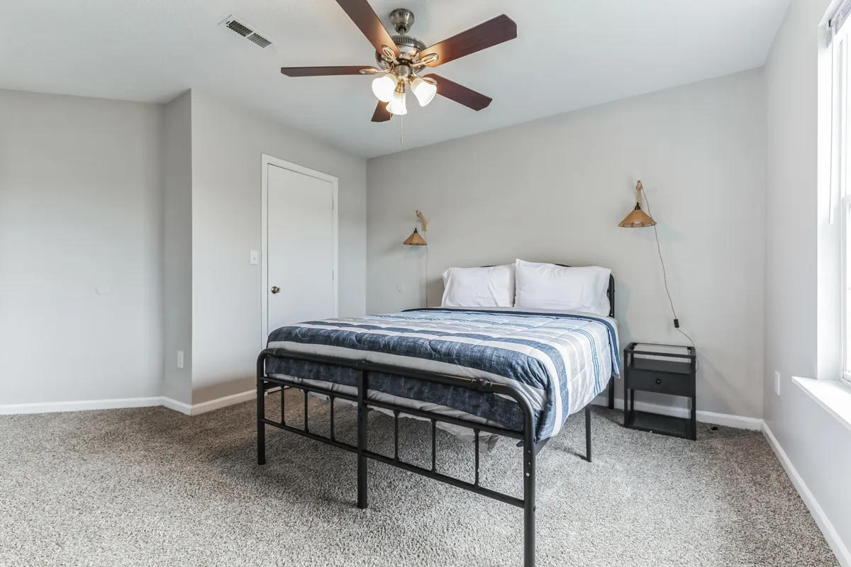 A cozy bedroom with blue striped bedding, ceiling fan, wall-mounted bedside lamps, and carpeted flooring. Sunlight streams into the room, creating a warm and inviting atmosphere.