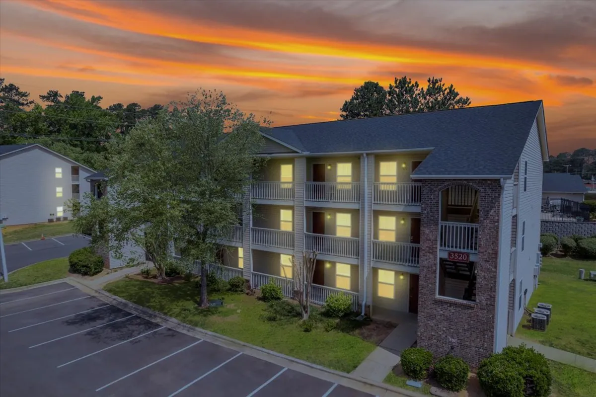 This image features the apartment building's facade bathed in the golden hues of sunset, with illuminated windows and residents enjoying their balconies. Some people are seen relaxing and socializing, while others take evening strolls in the parking area below.