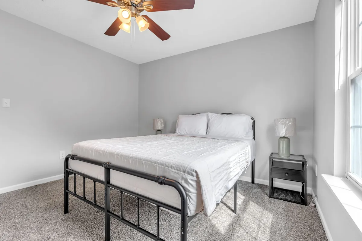 A tranquil bedroom with a comfortable queen bed, ceiling fan, nightstand, and warm bedside lighting. The space feels inviting, with an emphasis on relaxation and privacy.