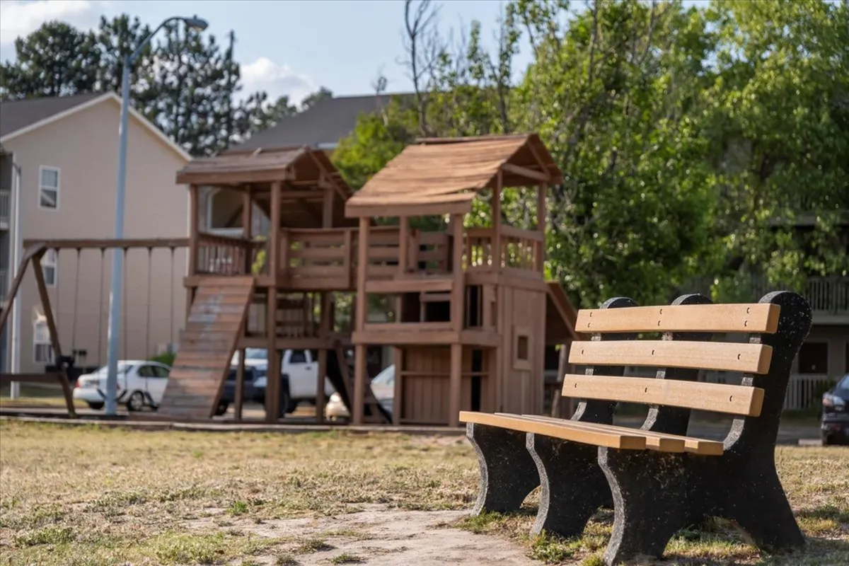 A welcoming playground structure sits beside a park bench, surrounded by trees and open space. Children are actively playing, while families watch and chat in the background, creating a joyful and inclusive atmosphere.