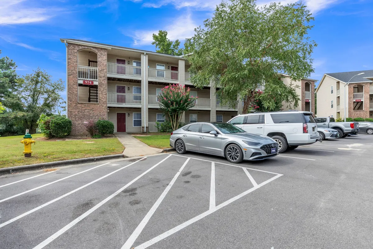 A bright exterior showcasing the apartment complex in daylight, with neatly landscaped grounds, parked cars, and residents partaking in leisure activities. The scene highlights sunny afternoons with people gathering in the courtyard and walking their pets.