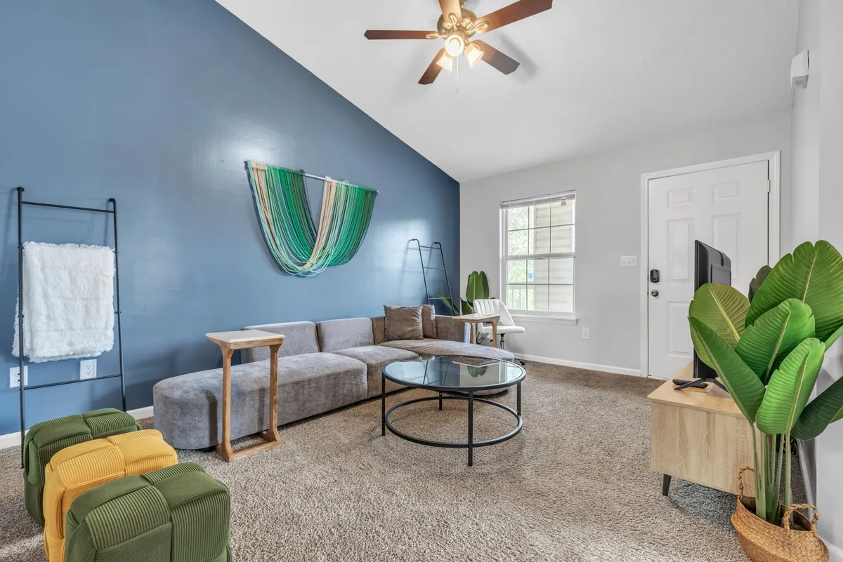 The image features a stylish living room with a bold blue accent wall, a comfortable sofa, a modern coffee table, eye-catching decorative wall art, and lush indoor plants. The scene exudes a casual and welcoming vibe, ideal for relaxing, hosting game nights, or enjoying a cozy movie marathon.