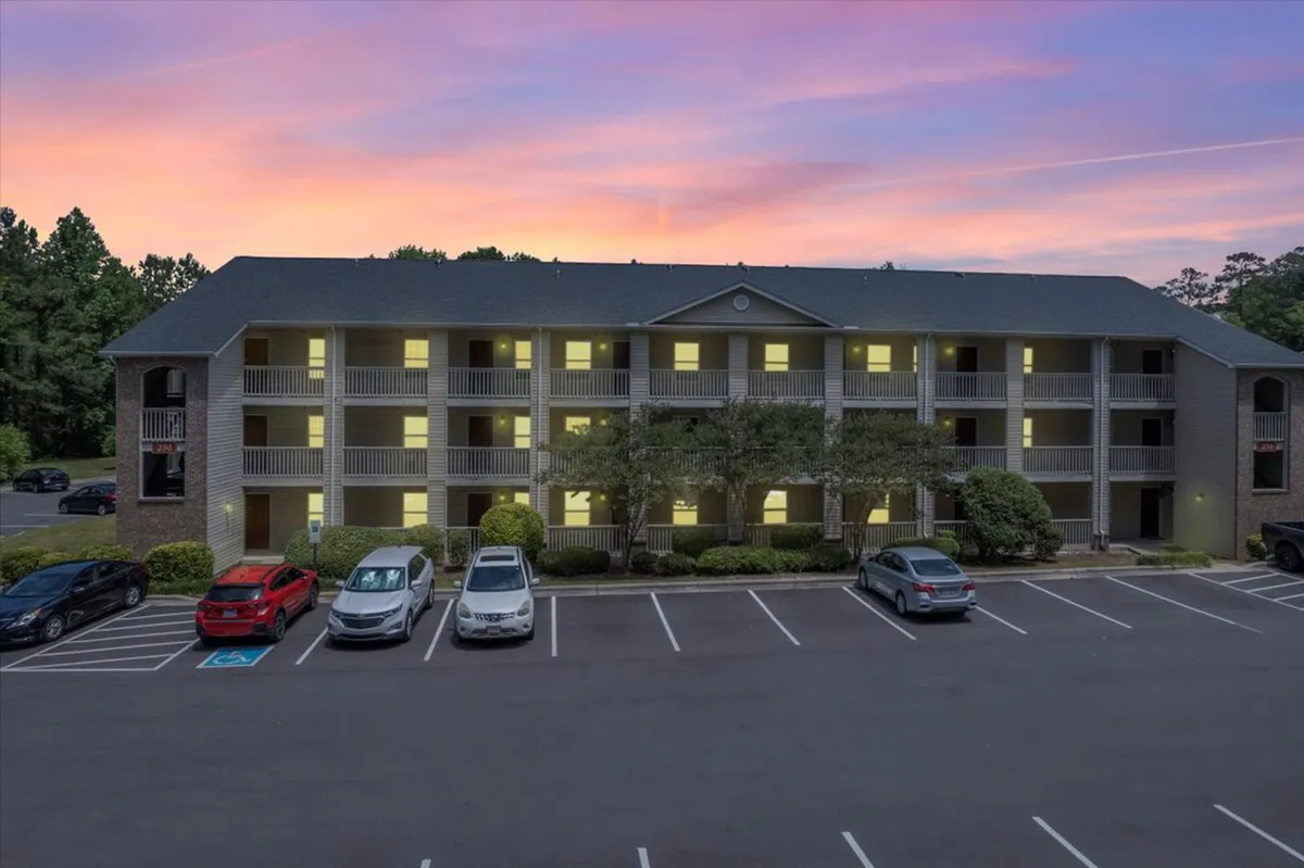 An apartment building with warmly illuminated windows during sunset, a visible parking lot, and a vibrant sky. Residents can be seen relaxing on balconies, hinting at a home-like atmosphere and the opportunity to unwind after a busy day.