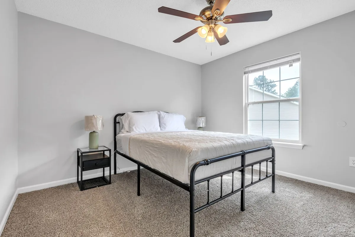 A peaceful bedroom scene featuring a neatly made bed, nightstand, lamp, and a bright window. The atmosphere is serene, suggesting comfort, relaxation, and the comforts of home.